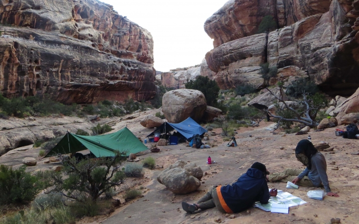 Between high canyon walls, people rest at a campsite with two tarp shelters. 